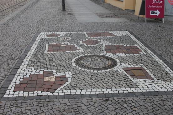 Großes, rechteckiges Mosaik, gelegt mit weißen und roten Steinen im Fußgängerzonenbereich.