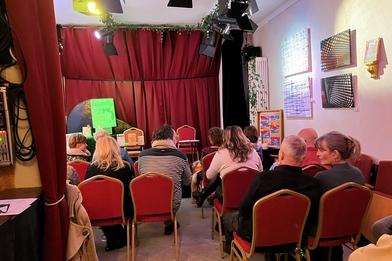 Foto der Kleinkusntbühne Lisa Zenner Art, Draufsicht Weinroter Theatervorhang, davor Bestuhlung mit Gästen..